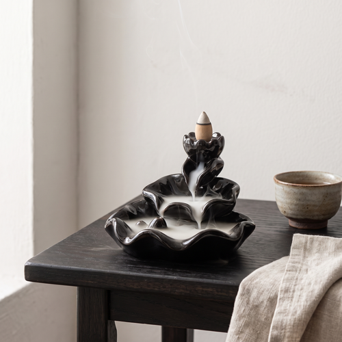 Black ceramic tiered lotus backflow incense burner with smoke cascading down, on a dark wooden tea table